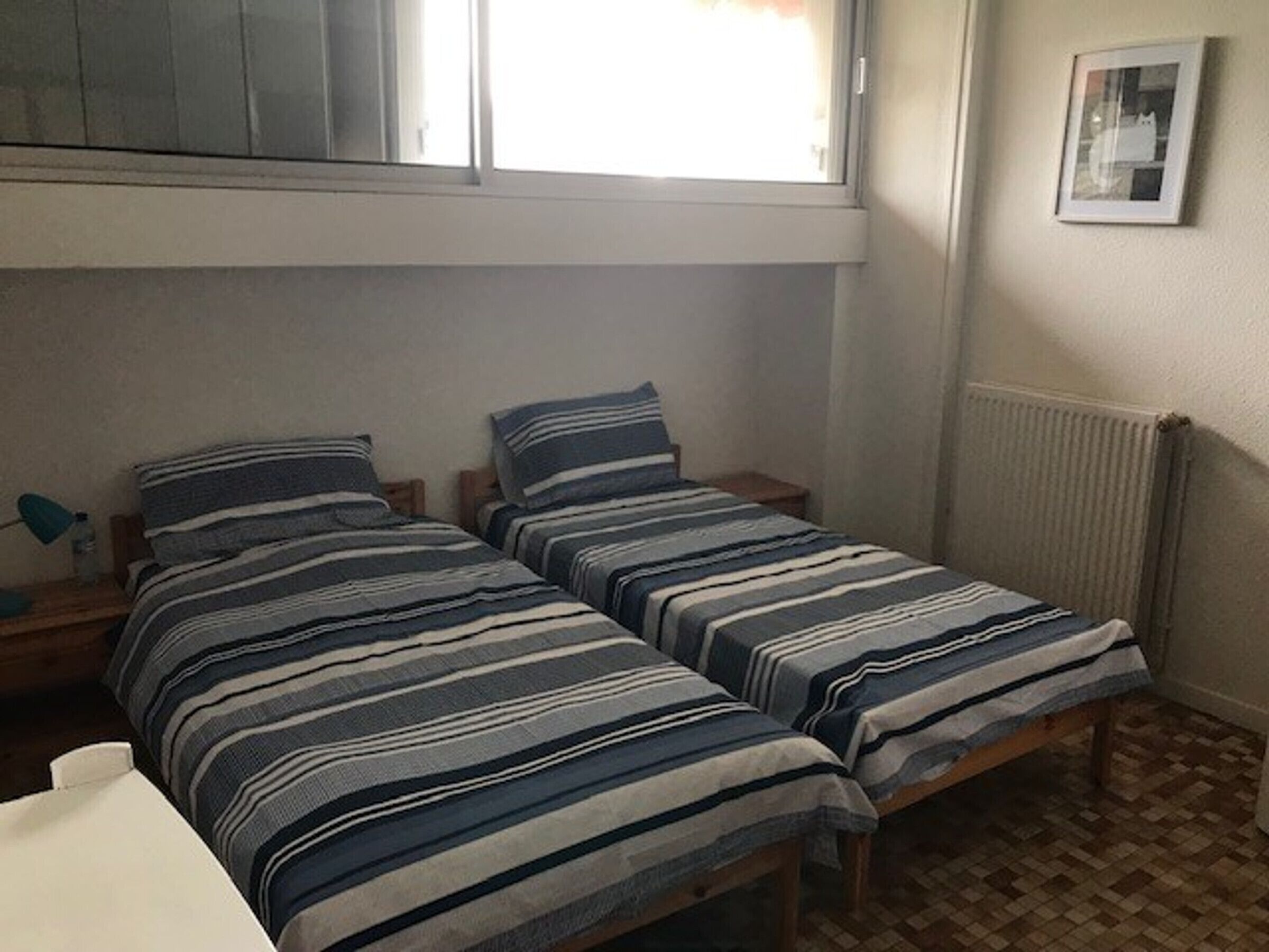 A small, single room with two single beds, a window, a radiator, and a framed picture on the wall.