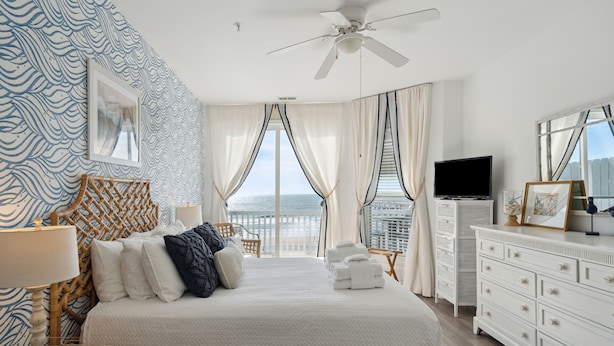 A hotel room with a large bed, a ceiling fan, a TV, a dresser, and a view of the city.