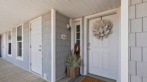 A covered porch with a wooden floor, a white door with a decorative wreath, and a small plant in a woven basket.