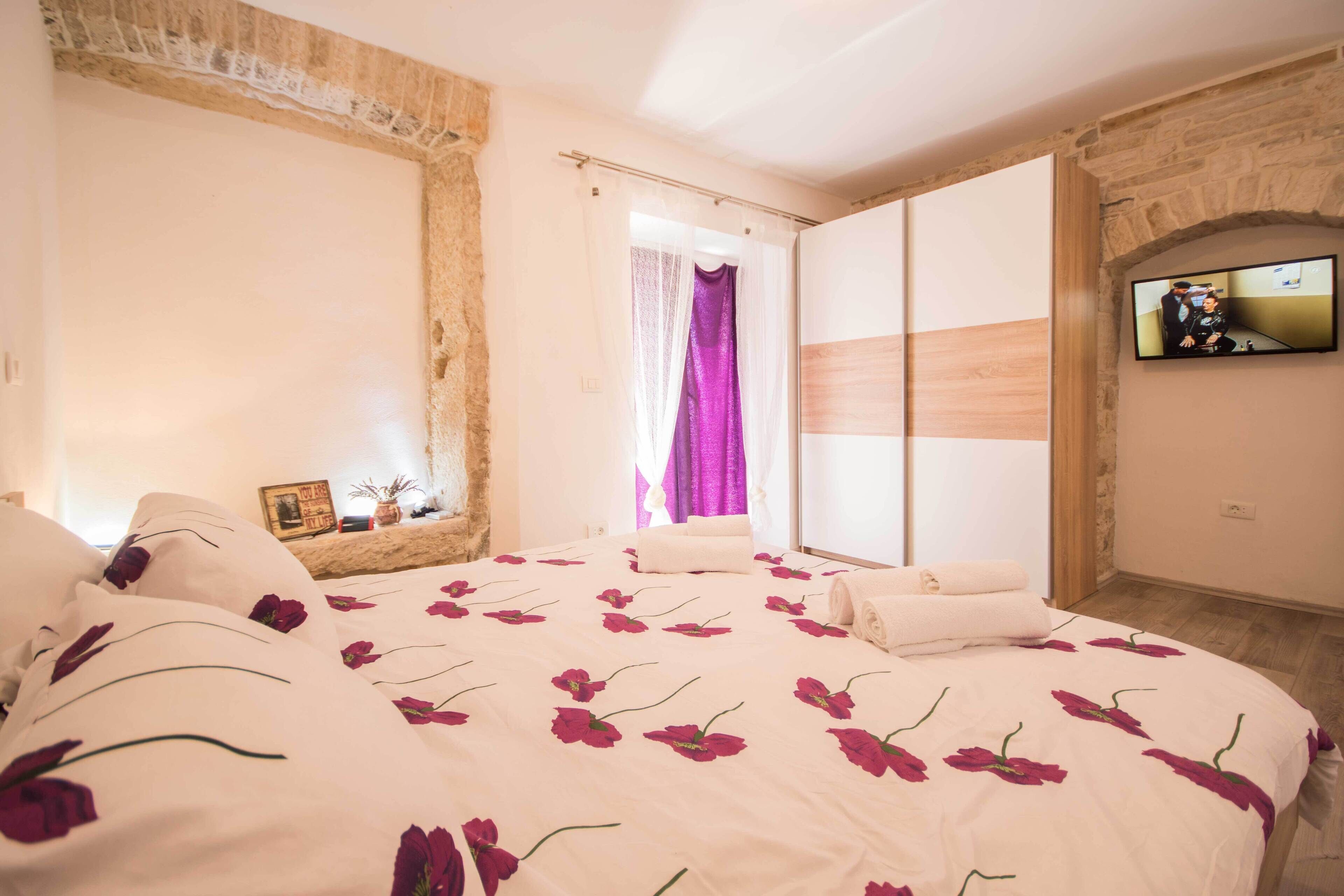 A bedroom with a large bed, a TV mounted on the wall, and a stone wall.