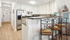 A kitchen with white cabinets, a stainless steel refrigerator, and a bar area with metal chairs. - Luxury Oceanfront with porch and swing daybed! Pet Friendly w/ fee, Elevator - Seaside Villa 101 (Folly Beach)