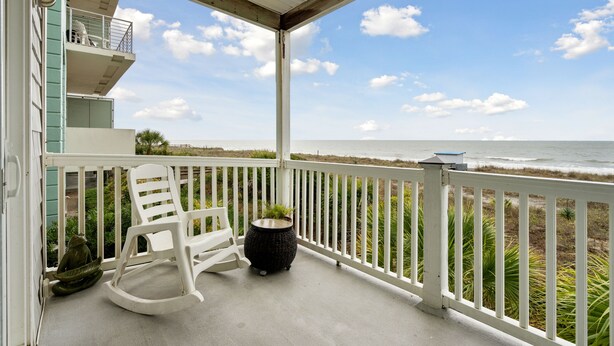 A balcony with a white railing, a white rocking chair, a small black table with a plant, and a view of the ocean. - Luxury Oceanfront with porch and swing daybed! Pet Friendly w/ fee, Elevator - Seaside Villa 101 (Folly Beach)
