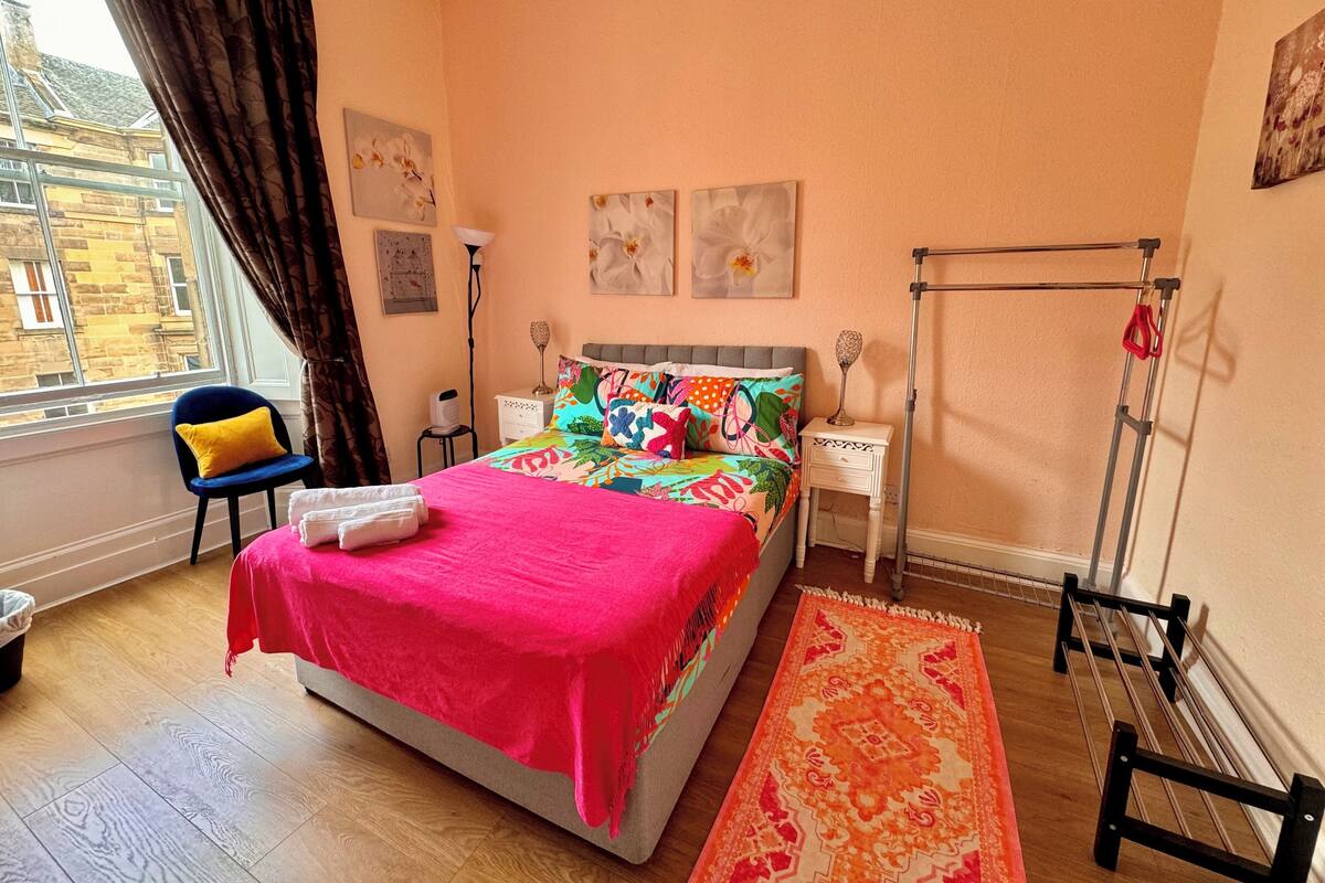 A bedroom with a bed, a chair, a nightstand, a lamp, a radiator, and artwork on the walls.