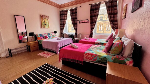 A well-lit bedroom with two beds, a large mirror, a wooden dresser, and a view of a building through the window. - @part Bernard Rooms (Edinburgh)