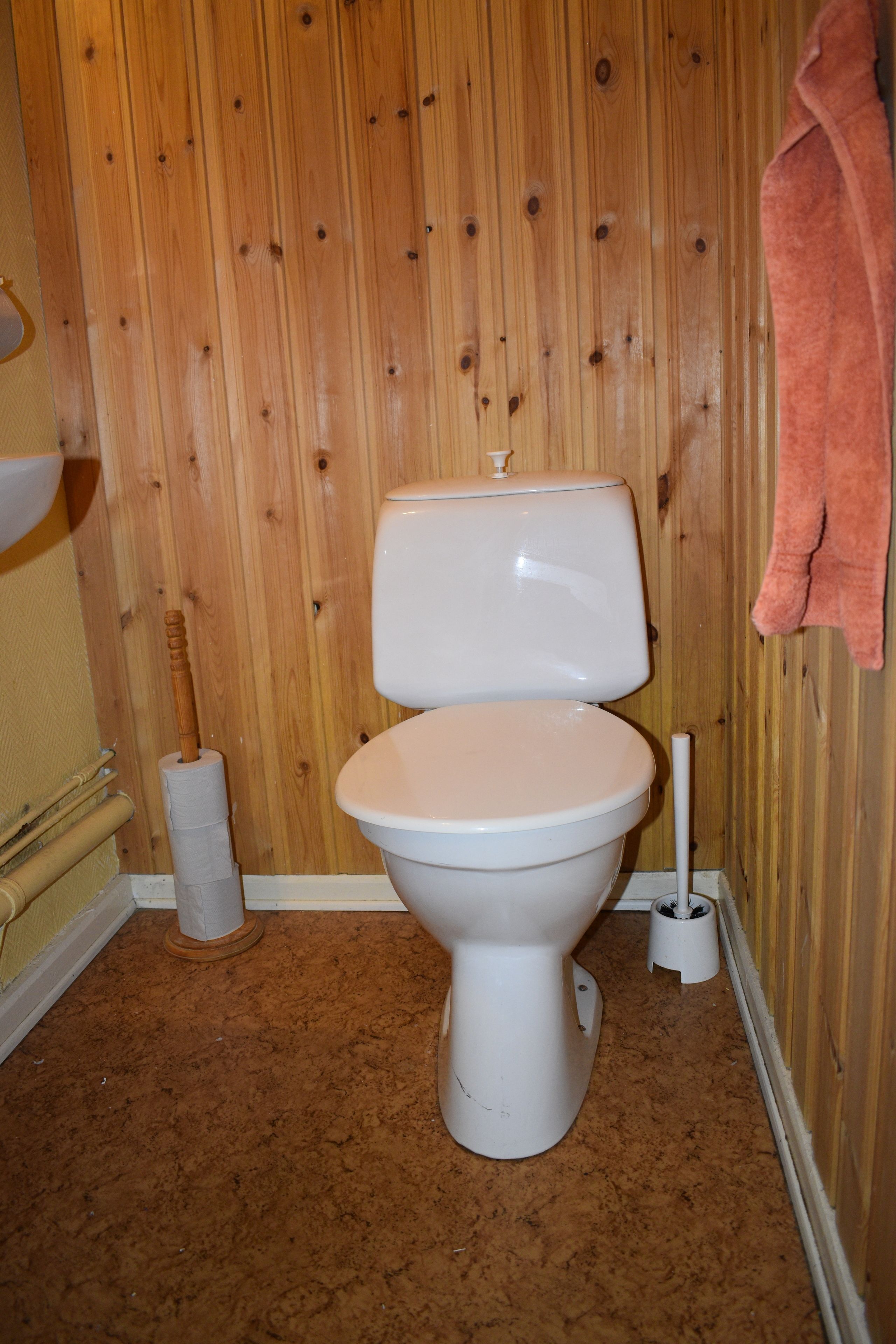 A bathroom with a wooden wall, a toilet, a roll of toilet paper, and a pink towel hanging on the wall.