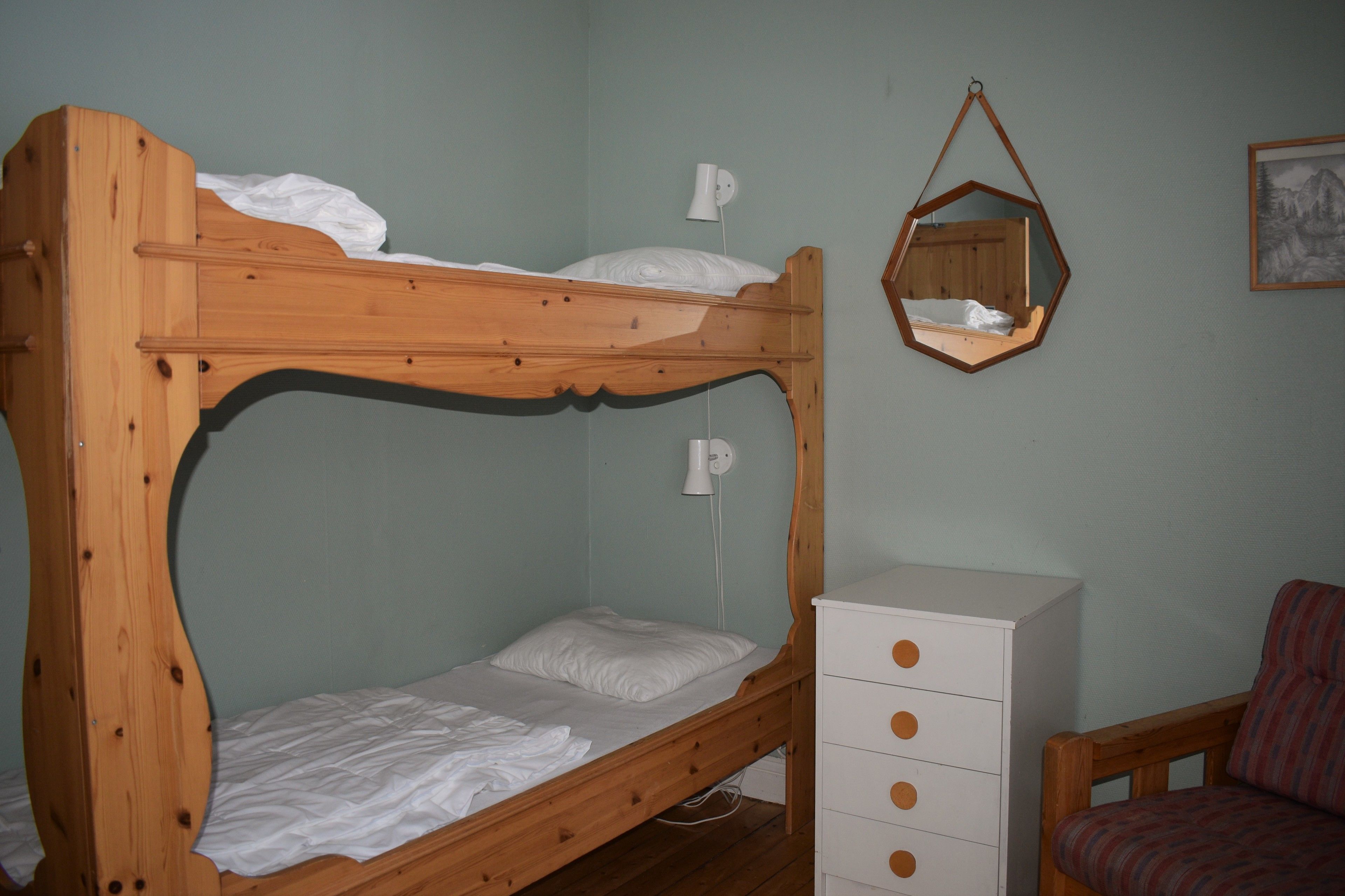 A bunk bed with a white mattress and pillows, a white dresser with drawers, a hanging mirror, and a framed picture on the wall.