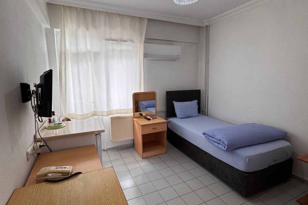A compact hotel room with a single bed, a desk with a chair, a small table, a mirror, and a television.