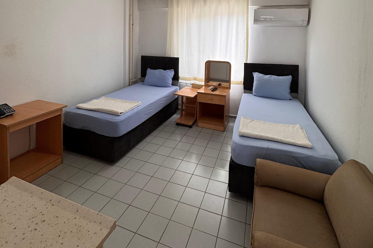 A hotel room with two single beds, a desk, a chair, a sofa, a ceiling light, and an air conditioning unit.