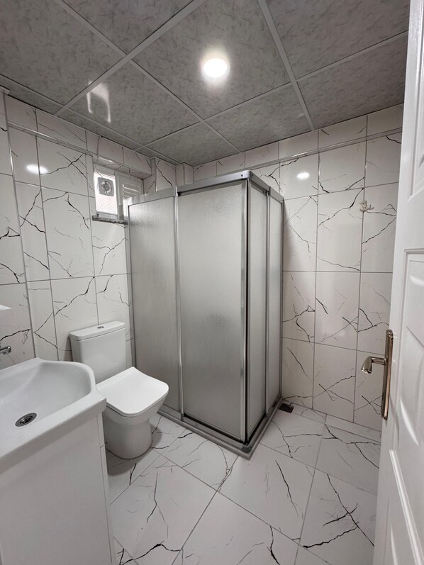A bathroom with white marble tiles, a glass shower enclosure, a white sink, and a toilet.
