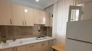 Una cocina con armarios color beige, encimera blanca, estufa a gas, lavaplatos y un refrigerador, además de un taburete pequeño.