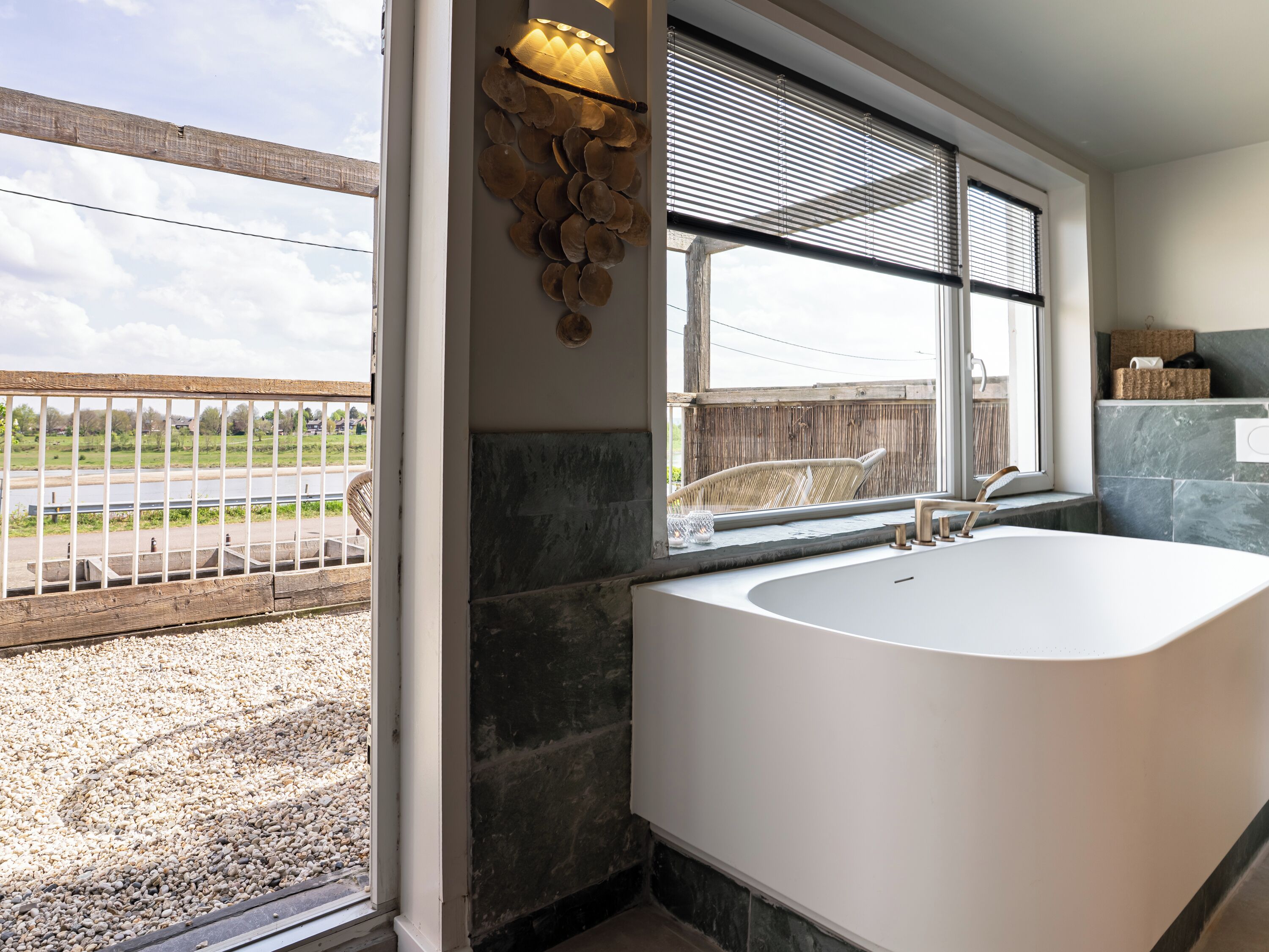 A bathroom with a large bathtub, a window with blinds, a wicker chair, and a view of a river and bridge.