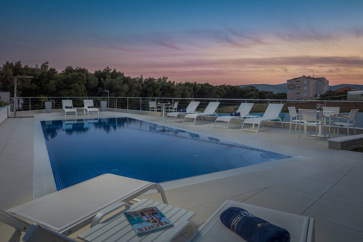 A rooftop pool area with lounge chairs and a table, surrounded by trees and a building in the background.