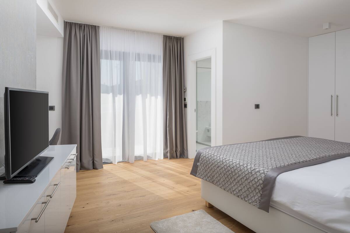A modern hotel room with a wooden floor, a bed with a patterned bedspread, a television on a white dresser, and a large window with sheer curtains.