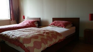 A double bed with a patterned bedspread, two bedside tables with lamps, and a window with curtains. - Les Tisserands (La Cambe)