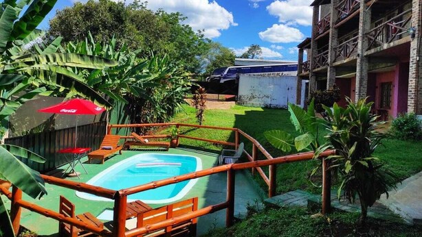 A pool area with a wooden deck and lounge chairs, surrounded by lush greenery and a building with a balcony. - Hotel Village del Sol (Puerto Iguazú)