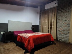 A bedroom with a bed, a nightstand, a lamp, an air conditioner, and a brick wall. - Hotel Village del Sol (Puerto Iguazú)
