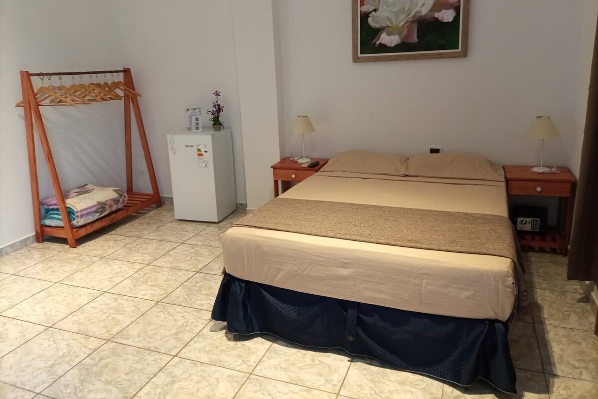 A bedroom with a bed, two bedside tables, a nightstand, a wooden wardrobe, a refrigerator, and a wall-mounted painting.