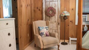 A room with a wooden wall, a white dresser, a beige armchair with a floral pillow, a floor lamp, and a small white stool. -  Cottage in the Woods- "Hummingbird"-- 2mi from Ricketts Glen falls trailhead! (Sweet Valley)