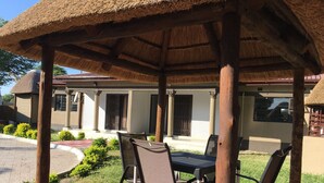 A covered outdoor seating area with a table and chairs, a thatched roof, and a building with columns in the background.