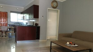 A modern living room with a sofa, coffee table, and a rug. A kitchen with cabinets and a clock is visible in the background.