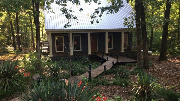 A rustic cabin with a metal roof, surrounded by a forest. It features a wooden deck leading to the entrance and is surrounded by various plants and trees. - Natchez Trace Cabin Retreat on 5 acres (Canton)