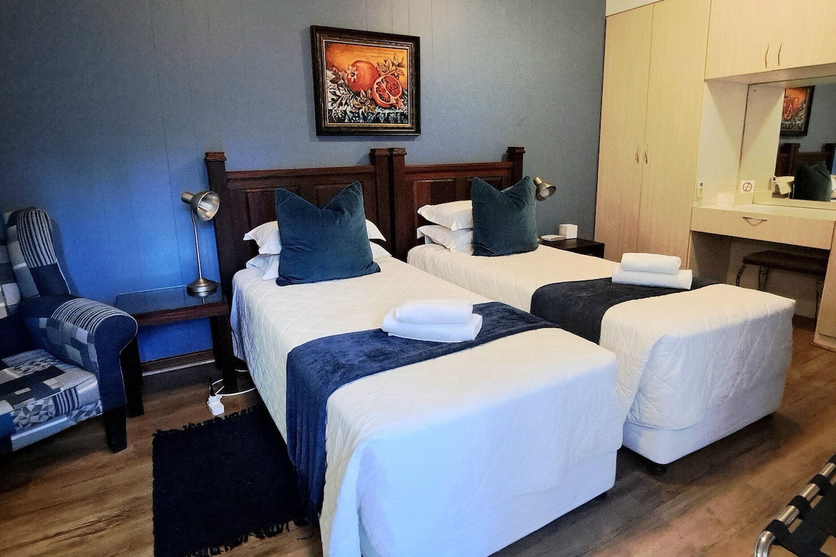 Ein Hotelzimmer mit zwei Einzelbetten, einem Holzkopfteil, einem Nachttisch mit Lampe, einem Sessel, einem kleinen Tisch und einem Spiegel.