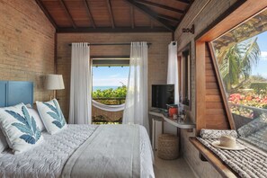 A bedroom with a large bed, a TV, a window with a view of the sea, and a balcony with plants. - Pousada Jeribá (Jijoca de Jericoacoara)