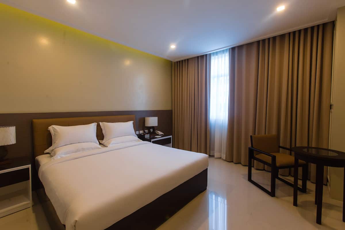 A hotel room with a large bed, a bedside table with a lamp, a chair, a small table, and a window with curtains.