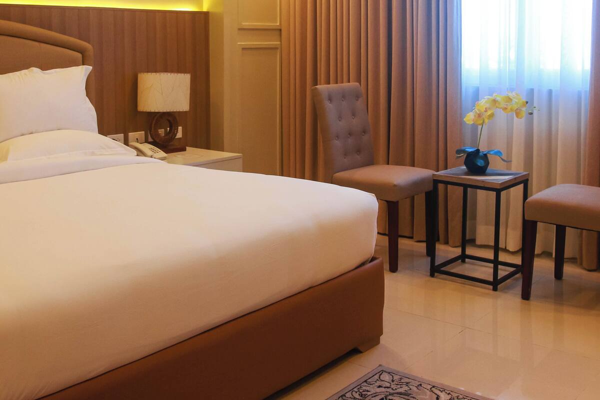 A hotel room with a large bed, a chair, a small table with a vase of flowers, and a nightstand with a lamp.