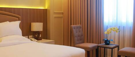 A hotel room with a large bed, a chair, a small table with a vase of flowers, and a nightstand with a lamp.