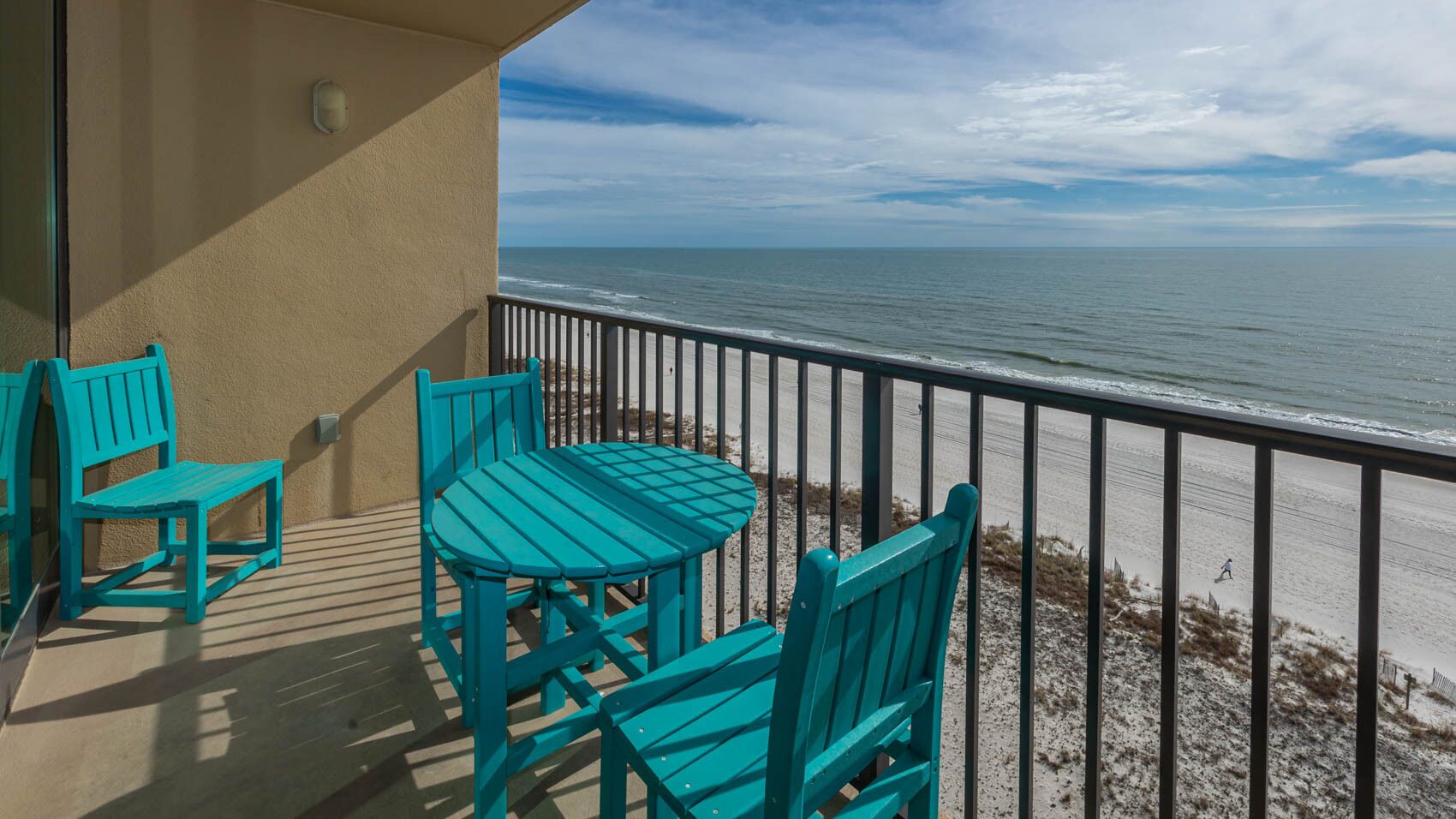 A balcony with turquoise chairs and a table, overlooking a beach with a railing.