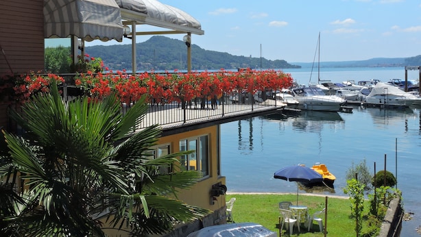 A waterfront property with a balcony overlooking a marina, a covered outdoor seating area, and a view of boats and hills in the distance. - Hotel Capri (Lesa)