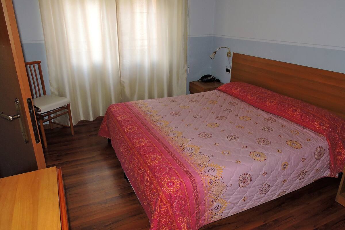 Quarto com cama de madeira, uma cadeira, mesa de cabeceira com abajur e janela coberta por cortinas.