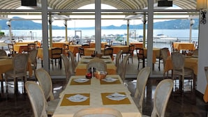 A restaurant with tables set for dining, large windows offering a view of the sea and boats, and a covered outdoor seating area. - Hotel Capri (Lesa)