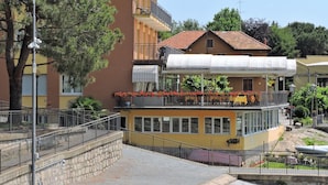 A multi-story building with balconies, a covered outdoor seating area, and a stone wall. - Hotel Capri (Lesa)