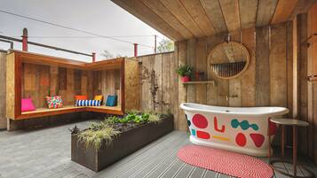 A wooden deck with a built-in bench, a large planter with plants, a freestanding bathtub with colorful designs, and a round mirror on the wall.