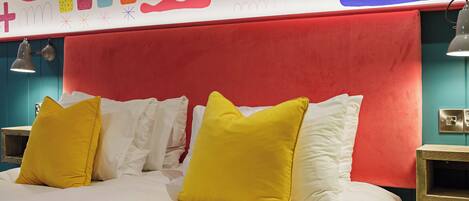 A bed with white and yellow pillows, a red headboard, and a colorful abstract wall design.