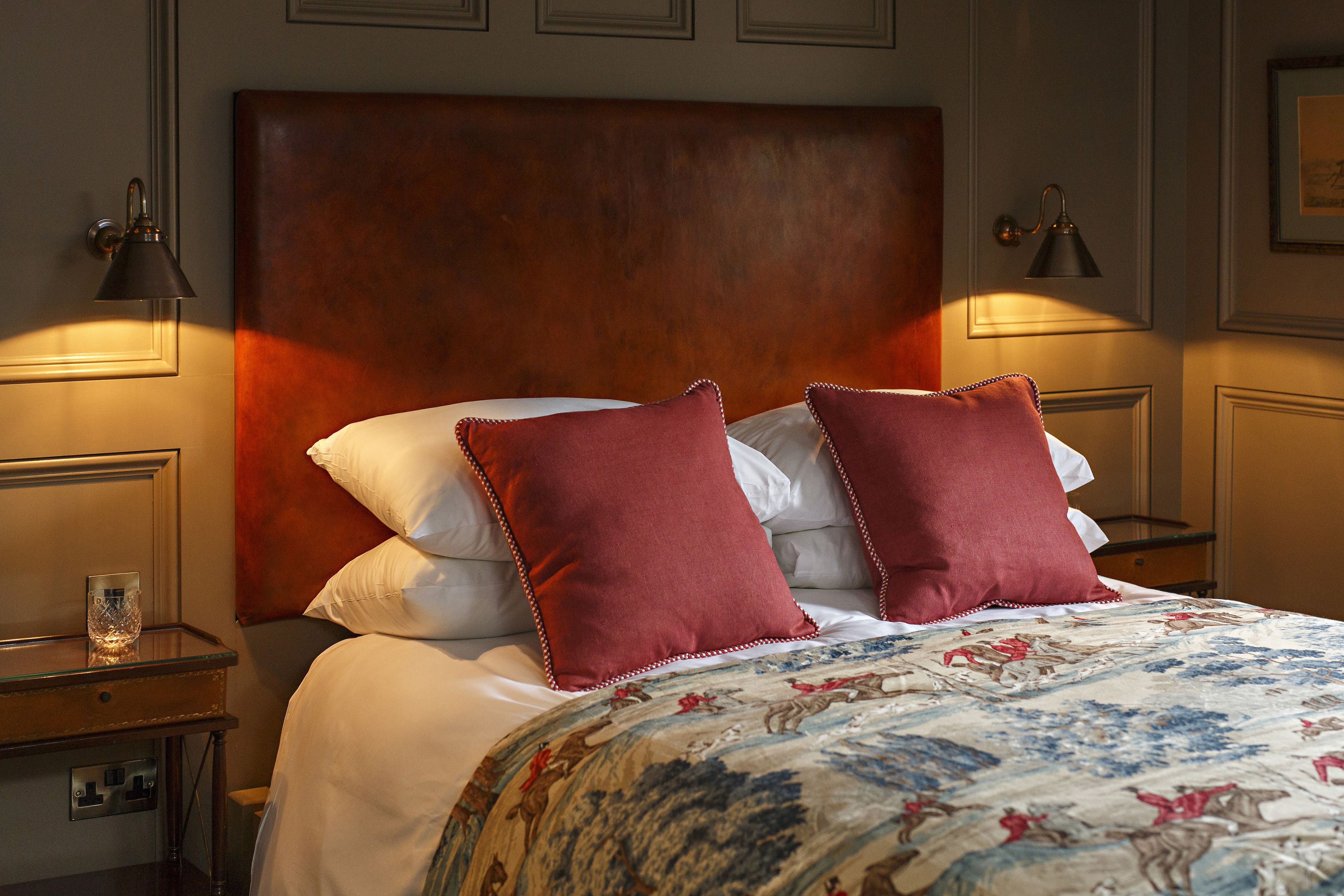 A neatly made bed with a floral bedspread and two red pillows, a wooden headboard, and two wall-mounted lamps on either side.