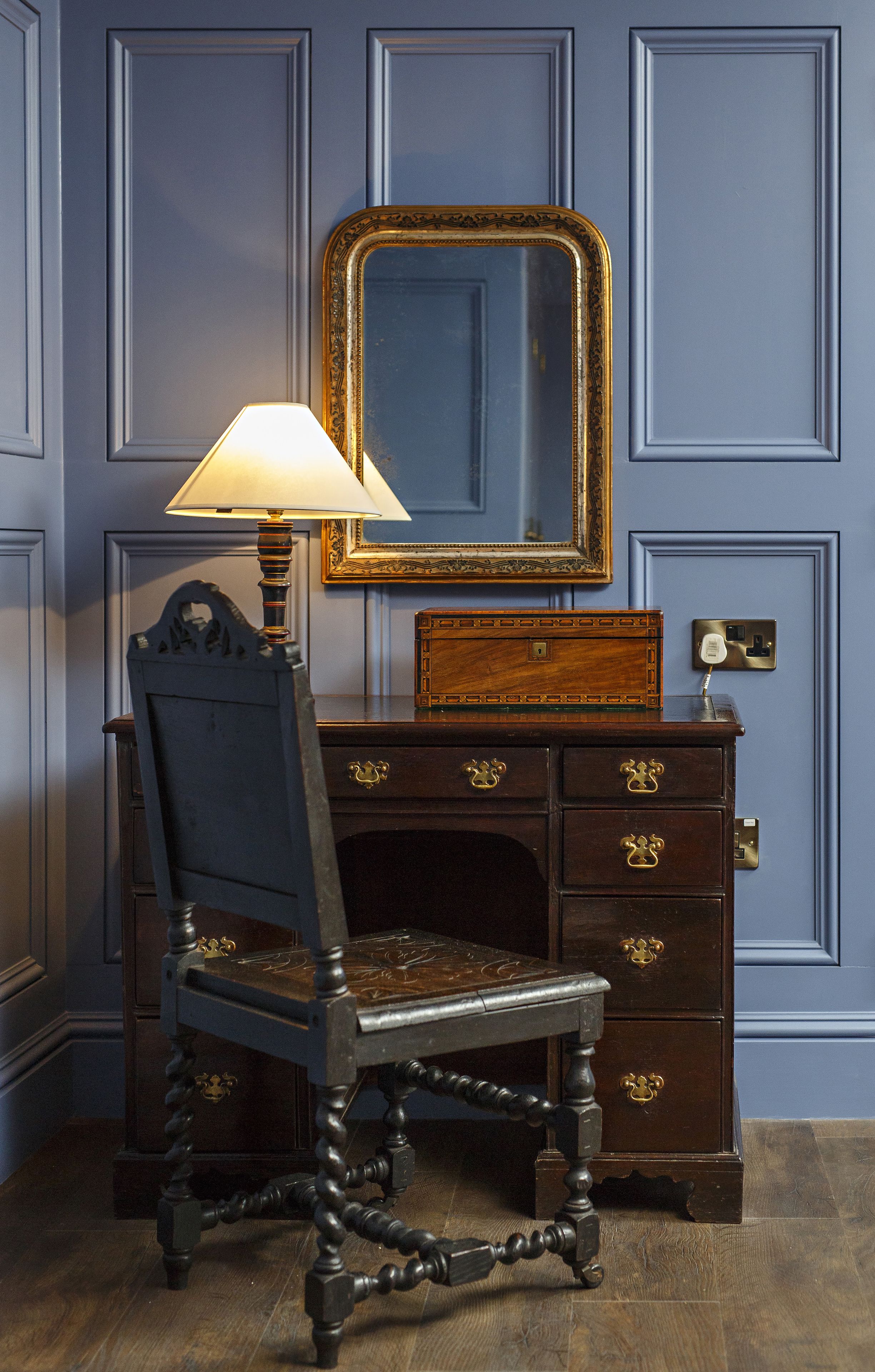 A dark wooden chair with a lamp, a mirror, and a wooden chest of drawers against a blue wall.