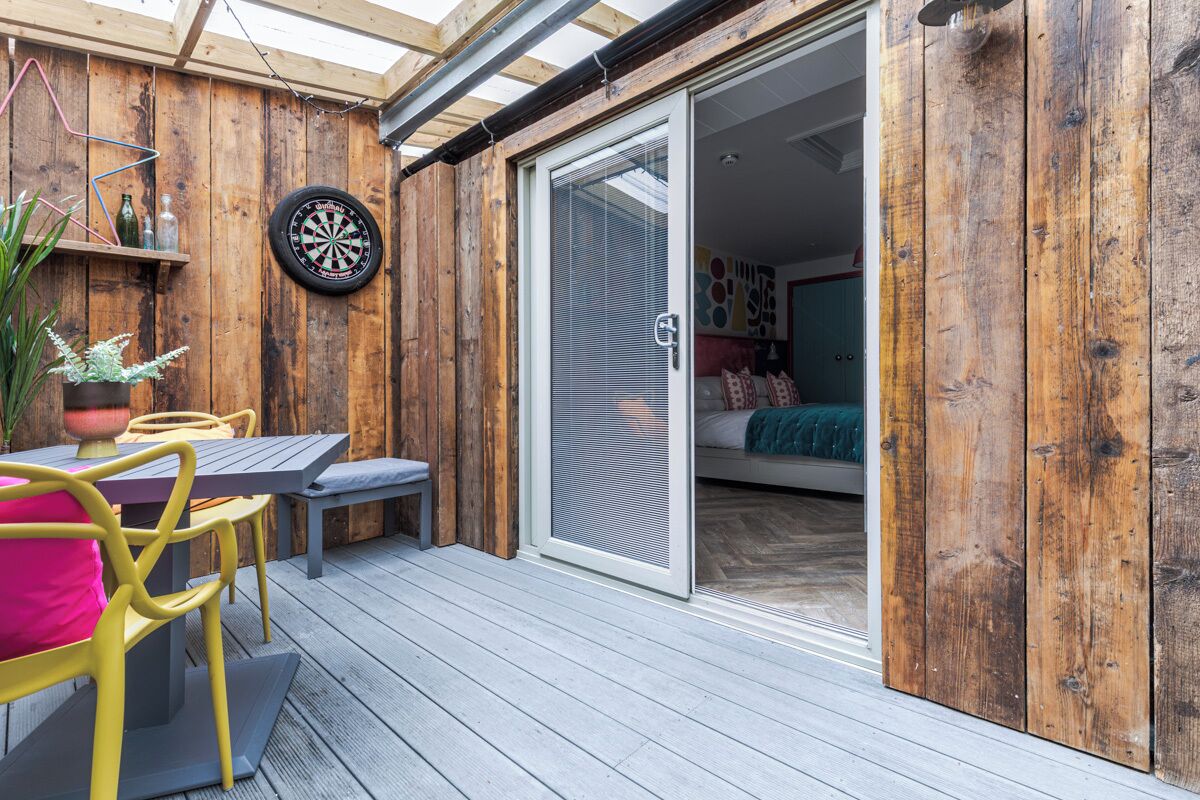 Une terrasse en bois avec une table et des chaises, un jeu de fléchettes et une porte-fenêtre coulissante donnant sur une chambre.