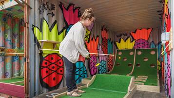 Une personne joue au minigolf dans une pièce colorée, décorée de graffitis, avec un gazon synthétique et des obstacles en bois.