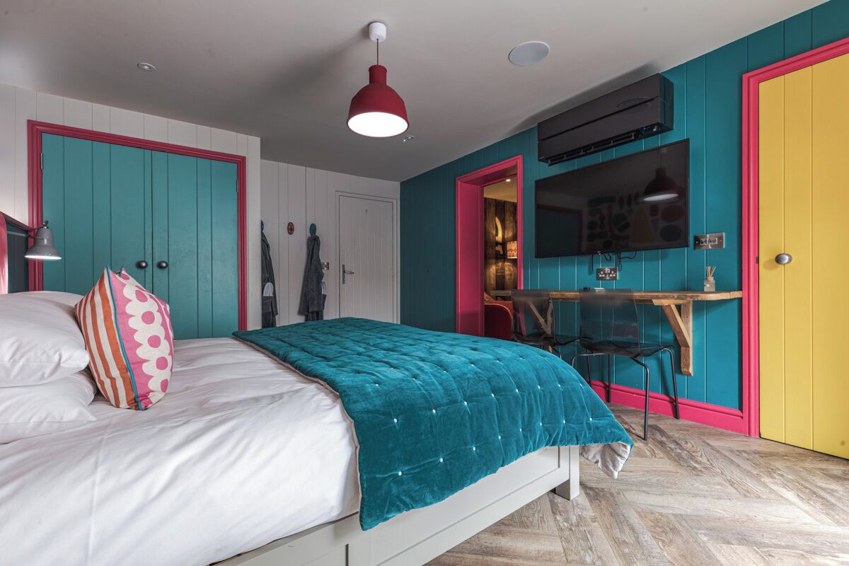 A bedroom with teal and pink walls, a bed with white bedding, a teal duvet, and a red pendant light.