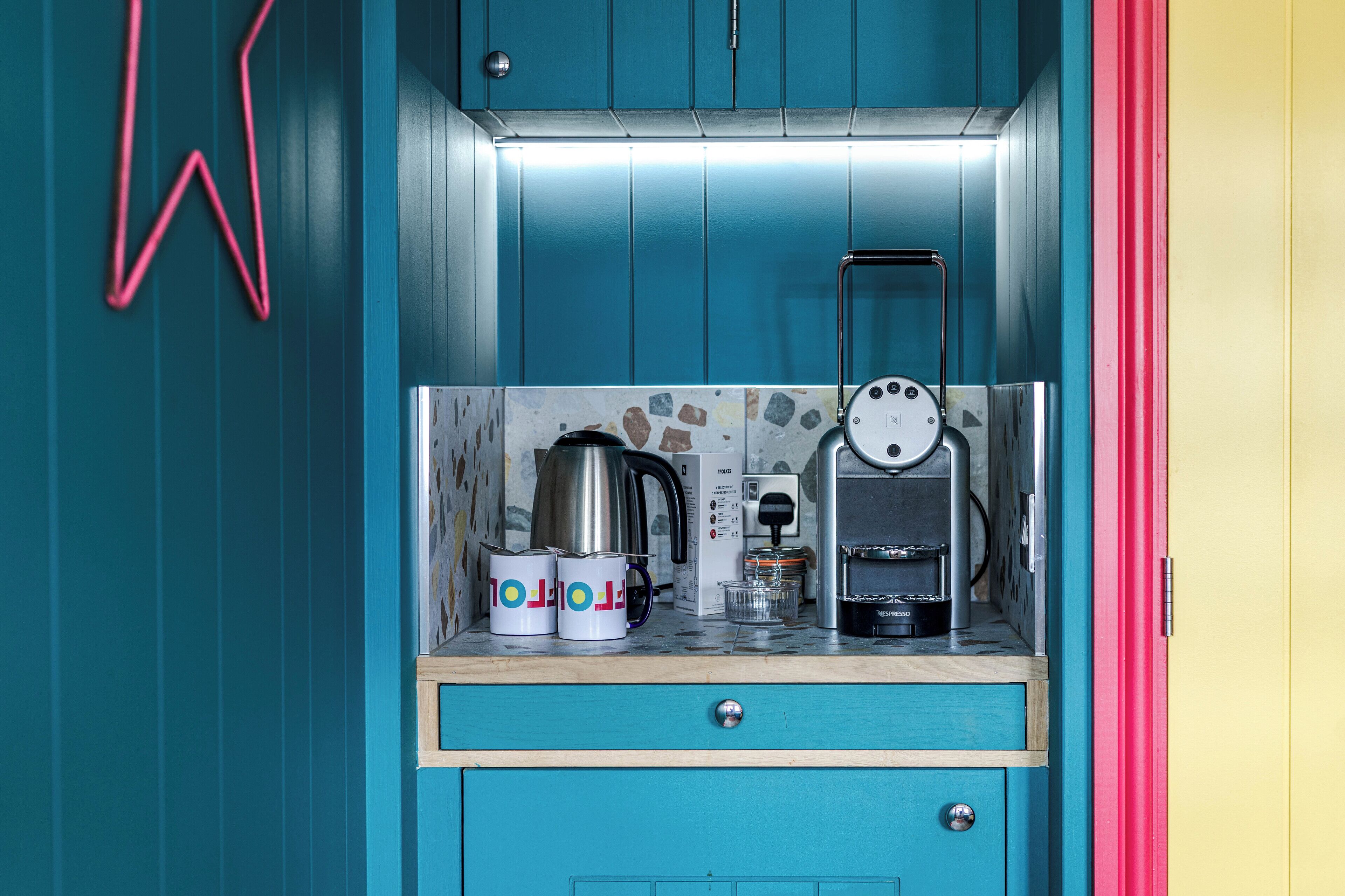 A kitchenette with a coffee machine, two mugs, a kettle, and a small cabinet.