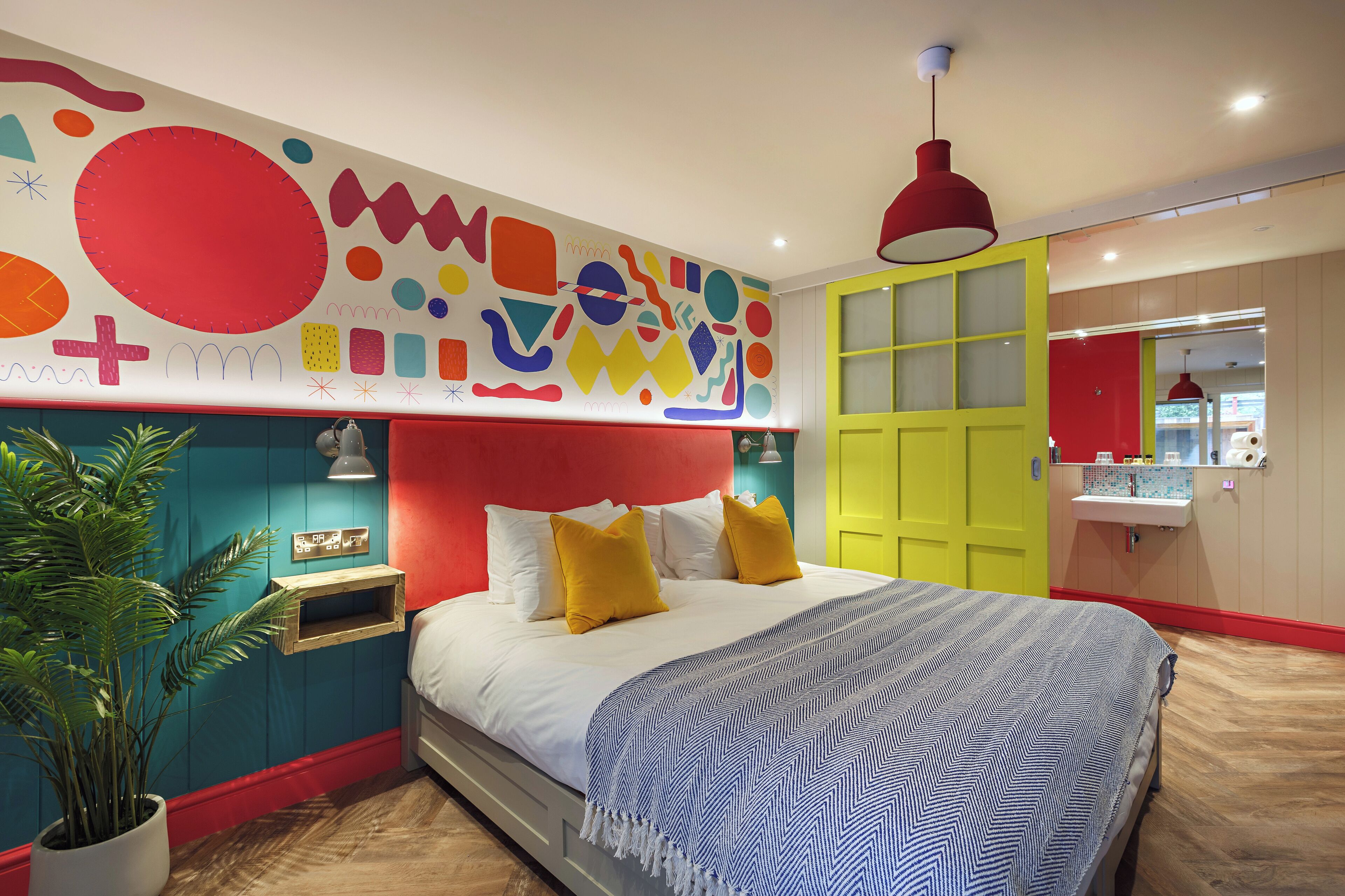 Une chambre moderne aux couleurs vives, avec un grand lit recouvert d’une literie à motifs, une fresque murale et une plante placée sur la table de chevet.