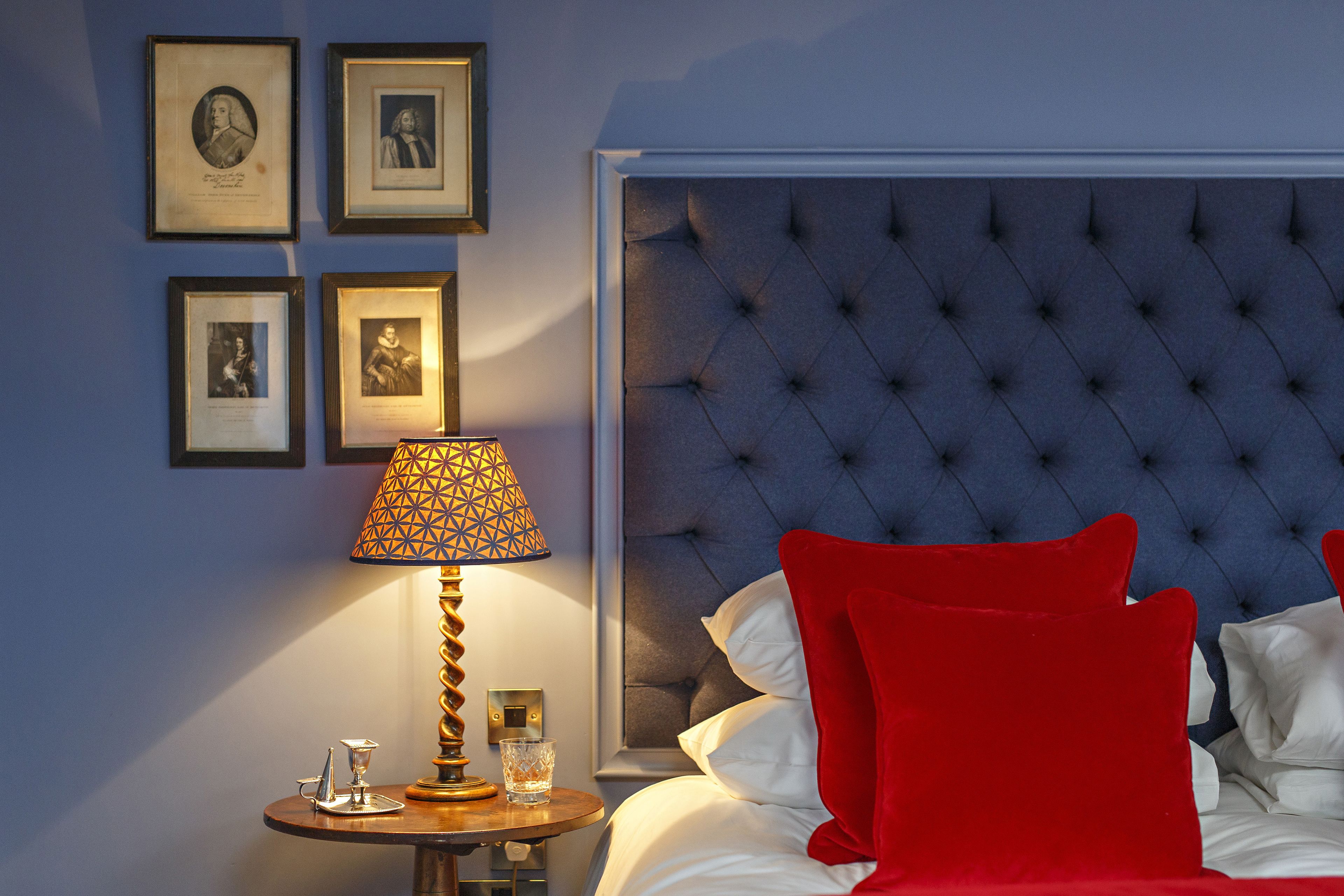A hotel room with a tufted headboard, a round bedside table with a lamp, and framed pictures on the wall.