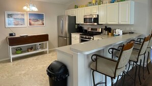 A modern kitchen with white cabinets, a stainless steel refrigerator, a microwave, and a gas stove. There are bar stools and a bench with a trash can.