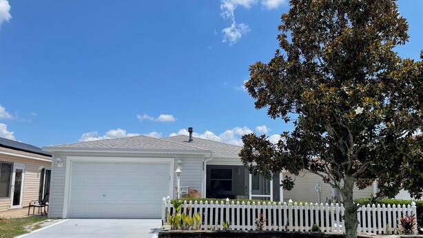 A single-story house with a garage, a white picket fence, and a tree in the front yard.