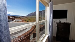 A room with a large window overlooking a road and mountains. There is a TV mounted on the wall and a small cabinet.