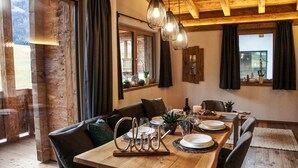 Een eetgedeelte met een houten tafel voor vier personen, omringd door donker lederen stoelen. Daarboven hangt een kroonluchter, en de kamer heeft zichtbare houten balken en grote ramen met gordijnen.