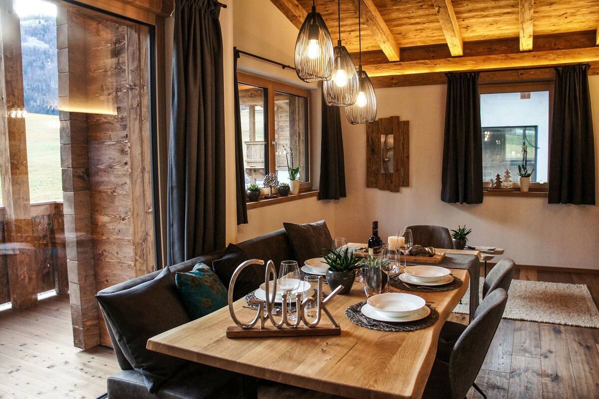 A dining area with a wooden table set for four, surrounded by dark leather chairs. A chandelier hangs above, and the room features exposed wooden beams and large windows with curtains.